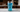 Ultra-detailed macro view of a tall glass containing nugget ice and electric blue liquid, positioned on a real surface with environmental context. The entire beverage system photographed as a unified physical object in real-world conditions.